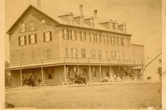 Historic sepia photograph of the Atlantic House, a large multi-story wooden hotel with wraparound porch and dormer windows, showing several people seated on the porch, photographed by McKenney of Portland, Maine.