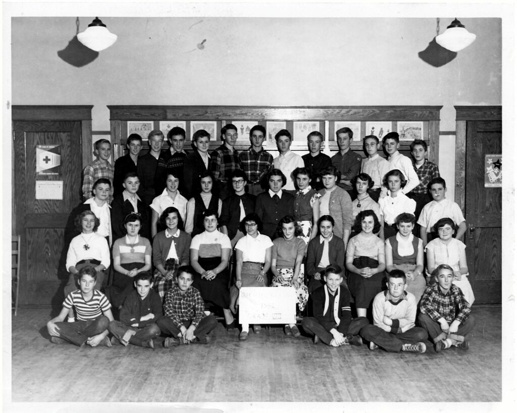 Group portrait of Oak Hill Grammar School Grade 8 students in Scarborough, Maine, 1952, posed in a classroom with boys standing and girls seated, with a class sign at center.